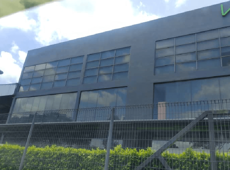 Modern gray office building with large glass windows, seen behind a metal security fence and trimmed green hedges under a partly cloudy sky