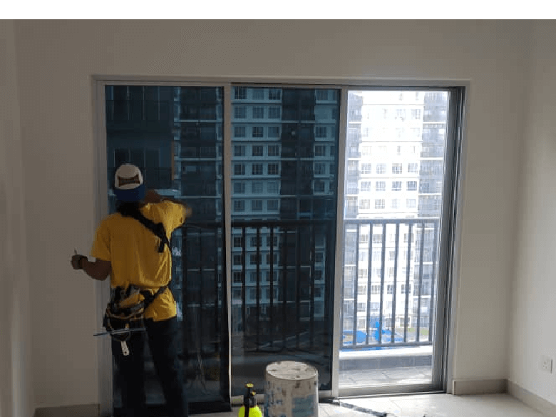 Worker in a yellow shirt and safety harness cleaning a sliding glass balcony door; bucket and spray bottle on the floor, city buildings visible outside.