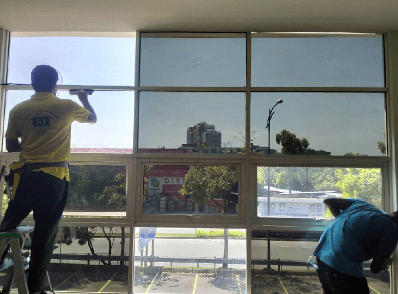 Two workers on a ladder and a platform are cleaning large office windows from inside the building with a street scene visible outside