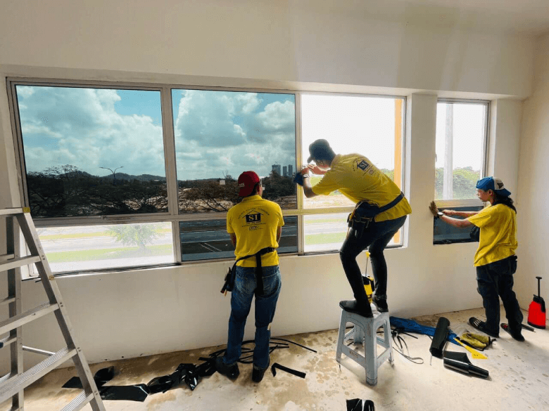Three construction workers in yellow shirts install large window panes in a bright room with tools on the floor and a ladder nearby