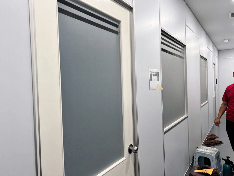 Long white-walled hallway with frosted-glass doors and metal door knobs; a person in a red shirt stands at the right edge, and a paper is taped to the wall beside a door.