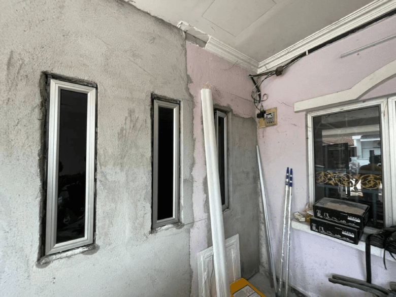 Interior of a renovation space with three tall narrow windows set in a gray plaster wall, pink wall behind, and scattered boards and tools.",